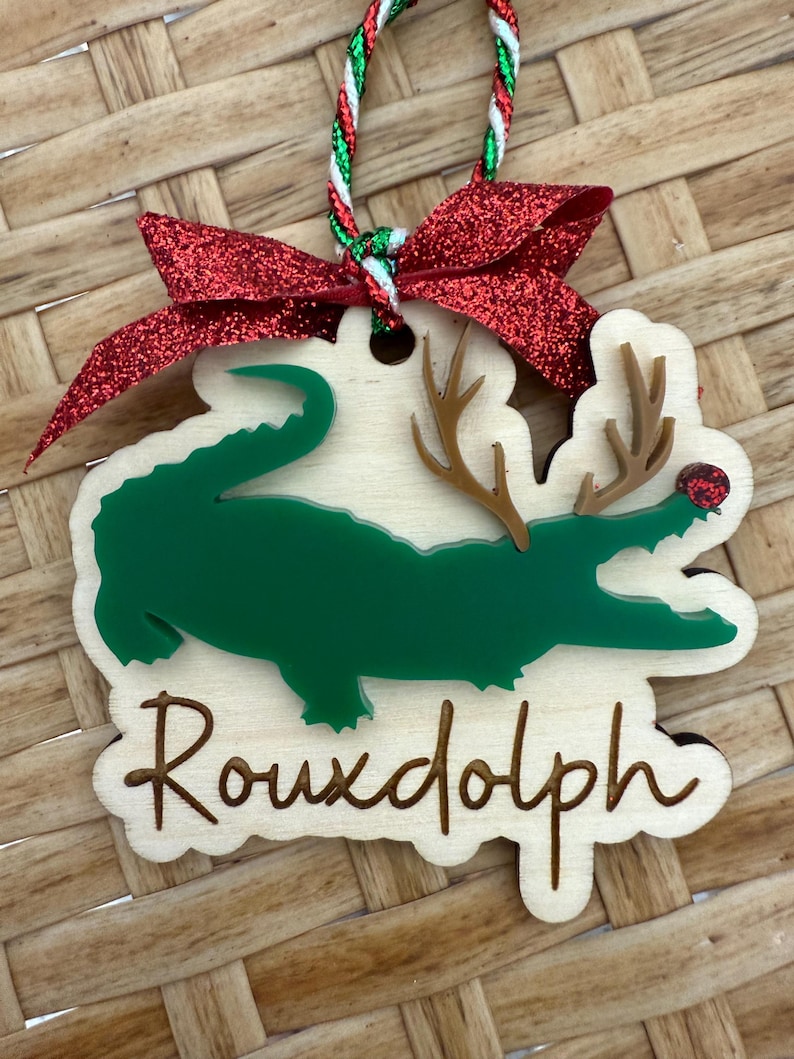 May include: Wooden Christmas ornament featuring a green alligator with antlers and a red nose, labeled "Rouxdolph." The ornament has a red glitter bow and a green, red, and white string for hanging. The background is a woven pattern.