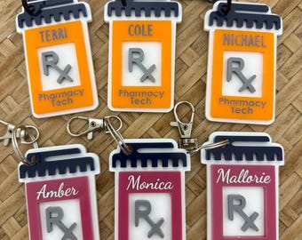May include: Keychains shaped like prescription pill bottles in orange and magenta, each with a name, the Rx symbol, and "Pharmacy Tech" printed on them. The keychains have black or silver hardware.