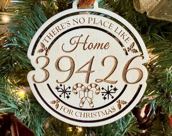 May include: Wooden Christmas ornament with the text "THERE'S NO PLACE LIKE Home 39426 FOR CHRISTMAS". The ornament has a brown ribbon for hanging and features snowflakes and candy cane designs. It is hanging on a Christmas tree.