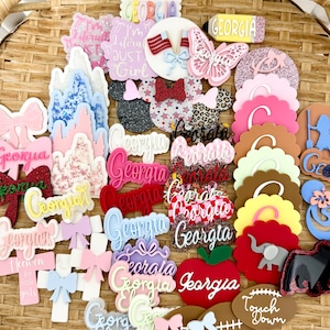 May include: A collection of assorted acrylic craft embellishments featuring various shapes including bows, castles, butterflies, apples, footballs, and mouse ears. Many pieces are personalized with the name Georgia, while others feature patterns like leopard print, glitter, and American flags.