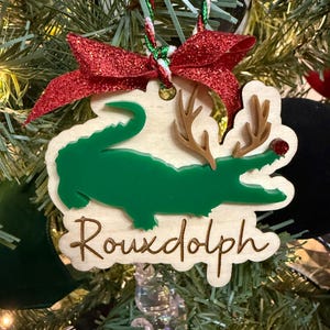 May include: A wooden Christmas ornament featuring a green alligator with brown antlers and a red nose. The ornament has the text "Rouxdolph" in a cursive font and is adorned with a red glitter bow.