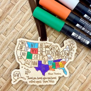 May include: A wooden ornament shaped like the United States map, with each state colored in. The ornament has a brown ribbon and the text "Travel far, travel often and travel without regrets - Oscar Wilde." Several colored markers are visible.