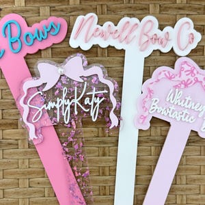 May include: Three custom acrylic display markers with long stems. One features a pink stem with teal text reading Ale's Bows. Another has a white stem with pink text reading Newell Bow Co. The third has a light pink stem with a bow design and text reading Simply Katy and Whitney's Bowtastic Bows.