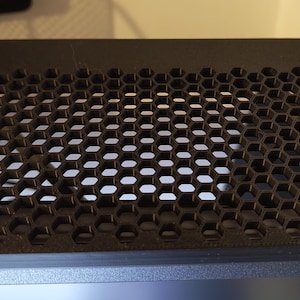 May include: A black, rectangular object with a honeycomb pattern. The object features numerous hexagonal apertures, forming a grid. It is constructed from a dark material, possibly a cover or screen, and is likely used for ventilation.