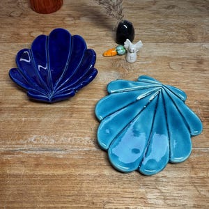 Op de afbeelding: Twee keramische schaaltjes in de vorm van een schelp, één in diep koningsblauw en de andere in helder turkoois. Beide schaaltjes hebben een glanzende afwerking en gegoten ribbels die vanuit de basis naar de geschulpte randen uitlopen.
