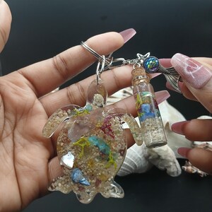 Könnte beinhalten: Ein klarer Schildkröten-Schlüsselanhänger aus Harz mit Sand, Muscheln und farbenfrohen Akzenten. Eine kleine Glasflasche mit Korken, gefüllt mit Sand und bunten Steinen, hängt neben der Schildkröte. Der Schlüsselanhänger hat einen silbernen Metallring.