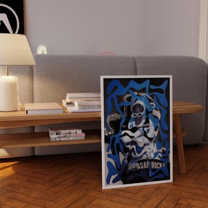 Op de afbeelding: Een ingelijste poster met een gestileerde afbeelding van A$AP Rocky in een wit en blauw ontwerp. De poster staat tegen een houten vloer, met een grijze bank en een houten bijzettafel met boeken en een lamp op de achtergrond.