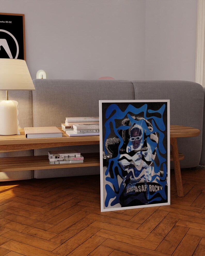 Op de afbeelding: Een ingelijste poster met een gestileerde afbeelding van A$AP Rocky in een wit en blauw ontwerp. De poster staat tegen een houten vloer, met een grijze bank en een houten bijzettafel met boeken en een lamp op de achtergrond.