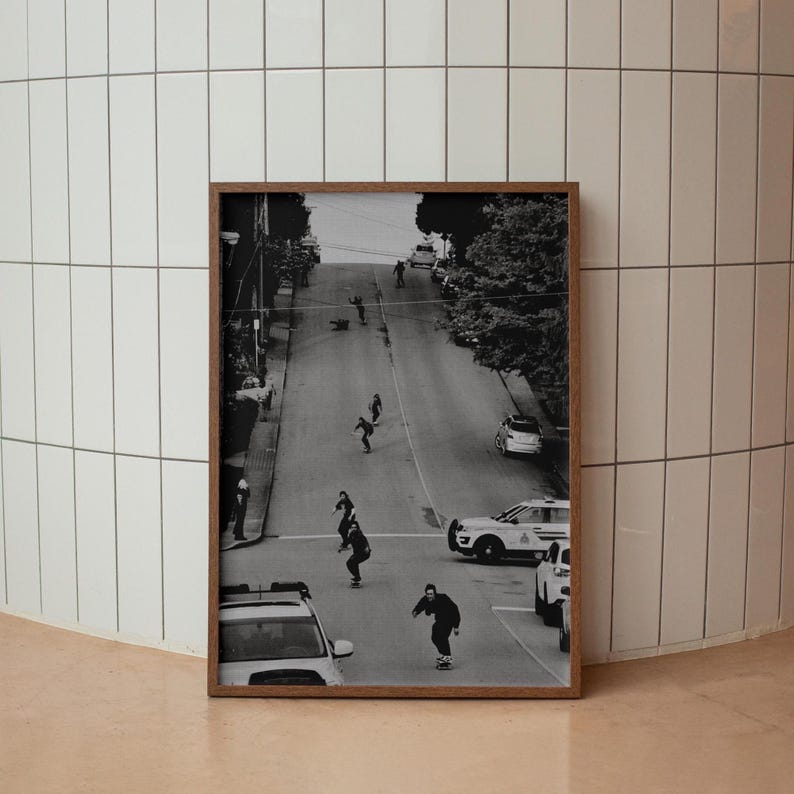 Op de afbeelding: Zwart-wit foto in een lijst van skateboarders die een steile straat afrijden. Auto's en bomen staan langs de weg. De afbeelding zit in een houten lijst.