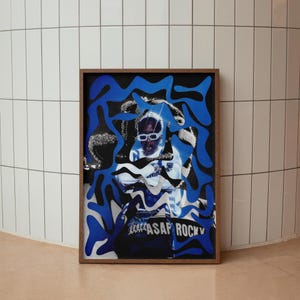 Op de afbeelding: Een ingelijste kunstdruk met een portret van een persoon met een zonnebril, overlegd met blauwe abstracte vormen. De print heeft de tekst "ASAP ROCKY" onderaan. De lijst is donkerbruin.