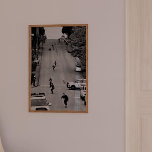 Op de afbeelding: Zwart-wit ingelijste foto van skateboarders op een stadsstraat. De afbeelding toont verschillende mensen die skaten op een lange, met bomen omzoomde weg, met auto's geparkeerd aan de zijkanten. De foto is gemonteerd in een lichtbruine houten lijst.