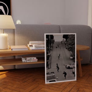 Op de afbeelding: Zwart-wit ingelijste foto van skateboarders op een straat. De afbeelding toont mensen die op een weg skaten, met auto's en gebouwen. De print staat op een houten vloer.