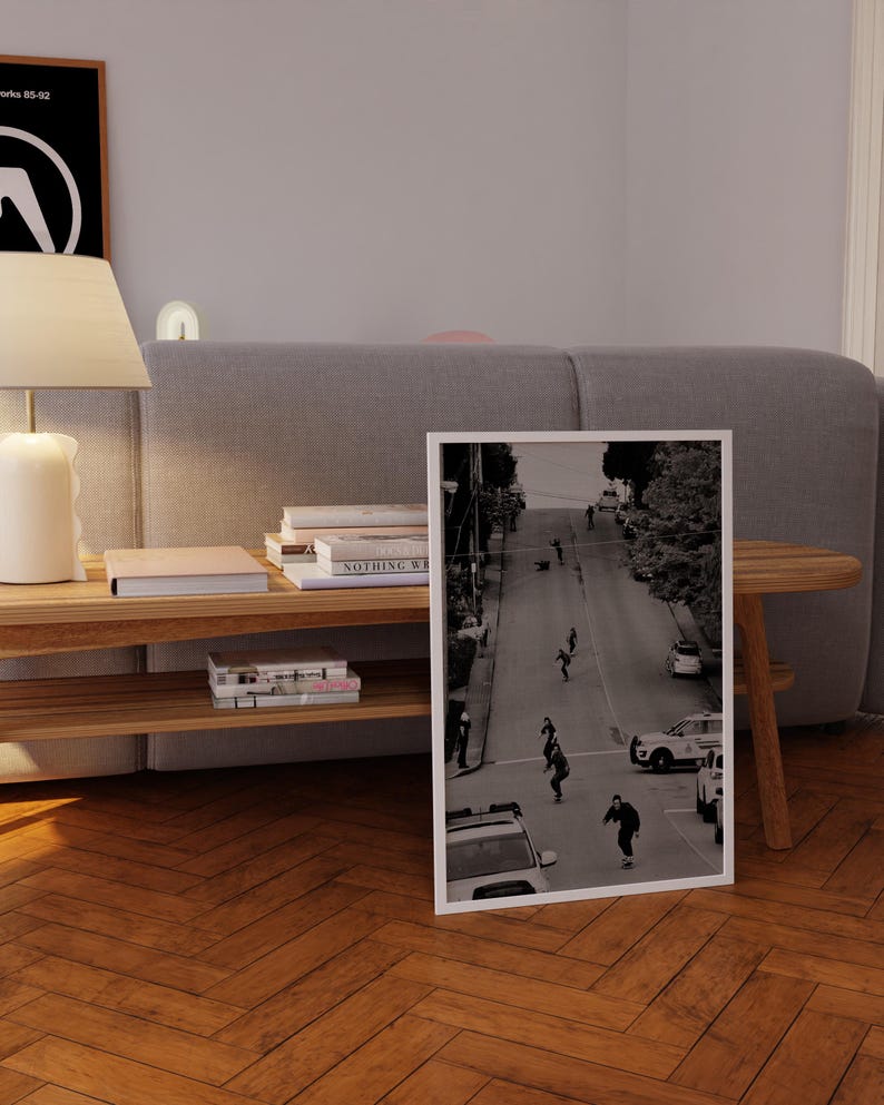 Op de afbeelding: Zwart-wit ingelijste foto van skateboarders op een straat. De afbeelding toont mensen die op een weg skaten, met auto's en gebouwen. De print staat op een houten vloer.