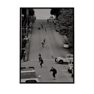 Op de afbeelding: Zwart-witfoto van skateboarders die een steile straat afrijden. Verschillende mensen skateboarden in het midden van de weg, met auto's geparkeerd aan de zijkanten. De foto is vanuit een hoge hoek genomen.
