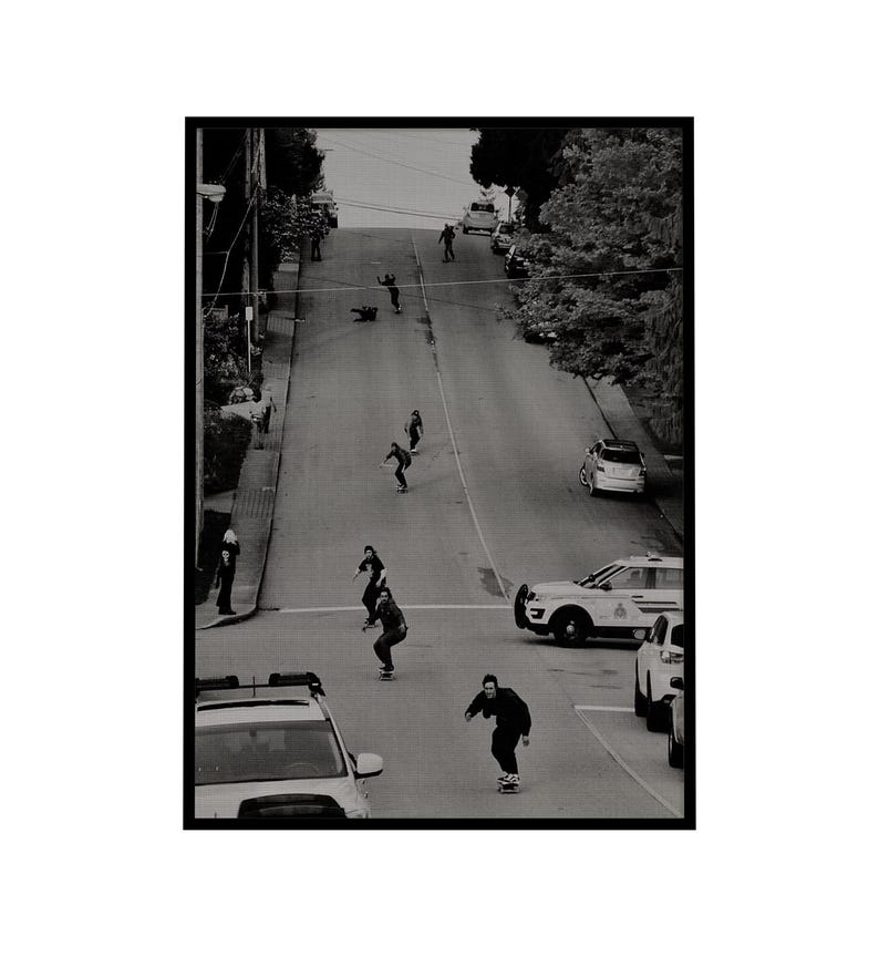 Op de afbeelding: Zwart-witfoto van skateboarders die een steile straat afrijden. Verschillende mensen skateboarden in het midden van de weg, met auto's geparkeerd aan de zijkanten. De foto is vanuit een hoge hoek genomen.