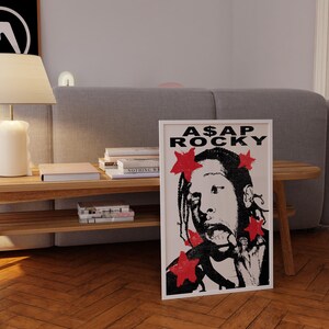 May include: A framed art print of A$AP Rocky in black and white, with red star accents. The print is on a wooden floor, propped against a grey sofa. The text "A$AP ROCKY" is at the top of the print.
