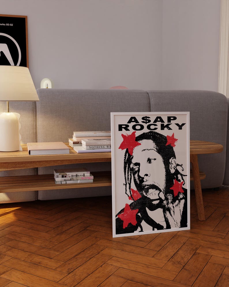 May include: A framed art print of A$AP Rocky in black and white, with red star accents. The print is on a wooden floor, propped against a grey sofa. The text "A$AP ROCKY" is at the top of the print.