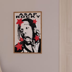 May include: A framed poster with a black and white illustration of A$AP Rocky, accented with red stars. The poster displays the text "A$AP ROCKY" in bold, black letters. The artwork is presented in a wooden frame.