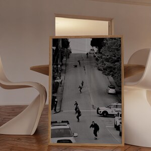 Op de afbeelding: Zwart-wit ingelijste print met skateboarders op een steile straat. De afbeelding toont verschillende mensen die een heuvel af skateboarden, met auto's en bomen langs de straat. De print wordt weergegeven in een kamer met een houten vloer en modern meubilair.