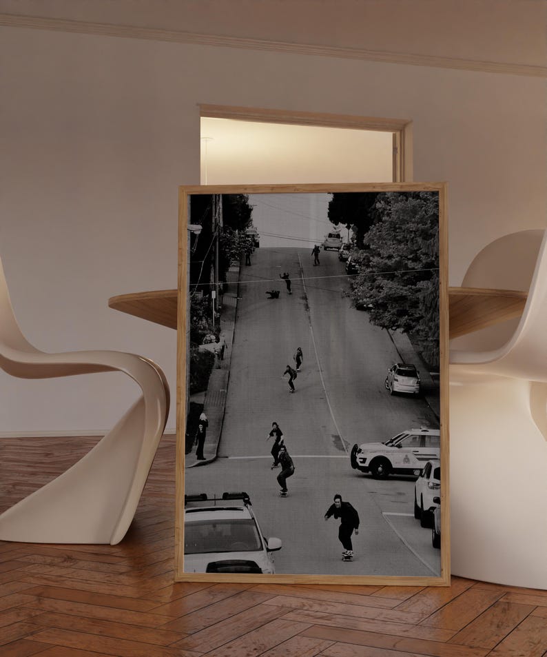 Op de afbeelding: Zwart-wit ingelijste print met skateboarders op een steile straat. De afbeelding toont verschillende mensen die een heuvel af skateboarden, met auto's en bomen langs de straat. De print wordt weergegeven in een kamer met een houten vloer en modern meubilair.
