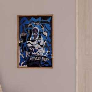 Op de afbeelding: Een ingelijste kunstdruk met een portret van een persoon met een zonnebril, overlegd met blauwe abstracte vormen. De print heeft de tekst "ASAP ROCKY" onderaan. De lijst heeft een natuurlijke houtkleur.