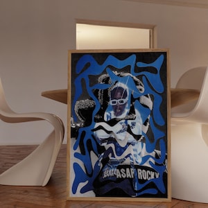 Op de afbeelding: Een ingelijste kunstdruk met een portret van een persoon met een zonnebril, bedekt met blauwe en zwarte abstracte vormen. De print heeft de tekst "ASAP ROCKY" onderaan. De lijst is van lichtgekleurd hout.