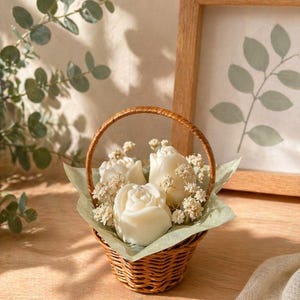 Può includere: Un piccolo cestino di vimini pieno di candele bianche a forma di rosa e delicati fiori bianchi. Il cestino ha un manico e poggia su un rivestimento di carta verde chiaro. Lo sfondo include una stampa botanica incorniciata e vegetazione.