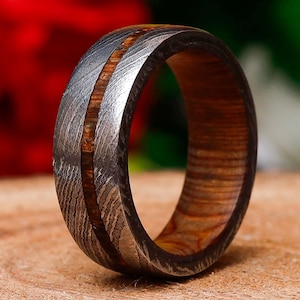 Può includere: Primo piano di un anello in acciaio Damasco con intarsio in legno. L'anello presenta un esterno in metallo scuro e strutturato con una fascia centrale in legno dai toni caldi. L'anello è posto su uno sfondo sfocato.