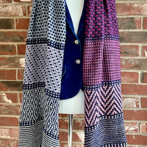 Op de afbeelding: Een blauwe en roze gestreepte gebreide sjaal met geometrische patronen. De sjaal is gedrapeerd over een mannequin die een blauwe blazer draagt.