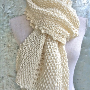 May include: Cream-colored, hand-knitted scarf displayed on a mannequin. The scarf features a chunky knit design with a scalloped edge. The soft, off-white color and textured pattern create a cozy and stylish accessory. Perfect for adding warmth and texture to any outfit.