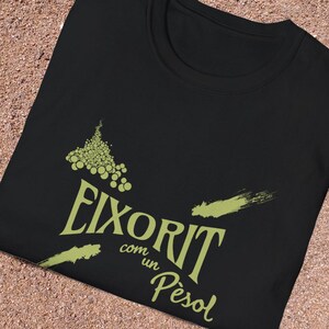 Puede incluir: Camiseta negra con un diseño verde de uvas y el texto "Eixorit com un Pèsol". El diseño incluye elementos similares a pinceladas. La camiseta está sobre una superficie arenosa.