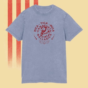 Puede incluir: Una camiseta azul claro con cuello redondo y mangas cortas. La camiseta presenta un gráfico circular rojo con una ilustración de gallo y el texto "XICA - CAMAGURTA - MICA - BALLARICA". El fondo tiene rayas rojas y blancas.