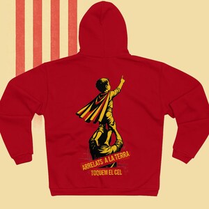 Puede incluir: Sudadera roja con capucha con un gráfico de un niño con capa sostenido por un adulto, señalando hacia arriba. El texto "ARRELATS A LA TERRA TOQUEM EL CEL" está debajo del gráfico. La sudadera tiene capucha.