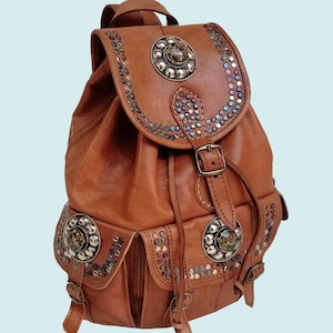 Può includere: Zaino in pelle marrone con borchie e abbellimenti argentati decorativi. Presenta una chiusura con patta, due tasche frontali e spallacci regolabili. Lo zaino ha una maniglia superiore e chiusure con fibbia.