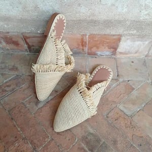 May include: A pair of woven raffia slip-on shoes. The shoes are beige with a pointed toe and a fringe detail across the top. The soles are brown. The shoes are displayed on a brick surface.
