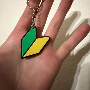 May include: A green and yellow beginner driver keychain with a black outline. The keychain is attached to a silver metal ring and chain. The design is a stylized Japanese beginner driver symbol.