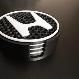 May include: A set of black coasters with a white Honda logo. The coasters have a honeycomb pattern and a white border. The set includes a black holder with a curved side for easy access to the coasters.