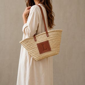 Peut inclure: Un sac fourre-tout en paille tressée naturelle avec des anses en cuir marron et un patch en cuir marron avec la lettre "L". Le sac est rectangulaire et porté à l'épaule.