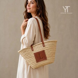May include: A woven straw tote bag featuring two brown leather shoulder straps and a square brown leather patch on the front centre. The patch is embossed with the capital letter L. The bag has a natural tan colour and a textured woven finish.