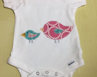 May include: White baby onesie with a pink and blue bird applique. The onesie has the text "Gerber Organic" and "onesies" on the tag.