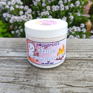 May include: A white jar with a pink and orange label featuring a cartoon cat and the words "paw print's toasted marshmallow fluff". The label also includes the text "DO NOT EAT". The jar is sitting on a weathered wooden surface.