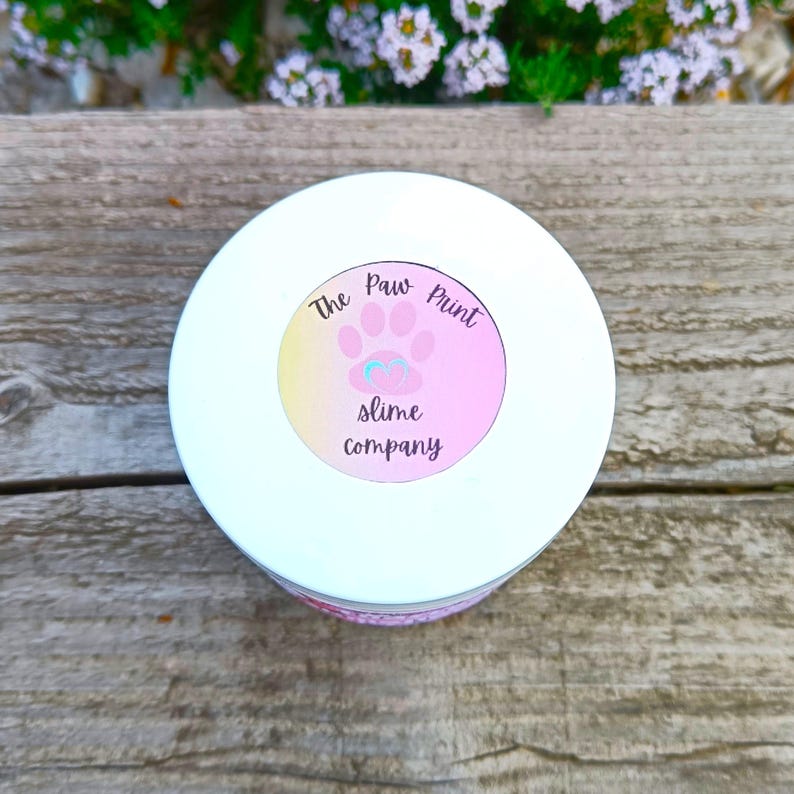 May include: A white container with a circular label featuring a pink paw print and the text "The Paw Print slime company." The label has a gradient of yellow to pink. The container sits on a wooden surface.