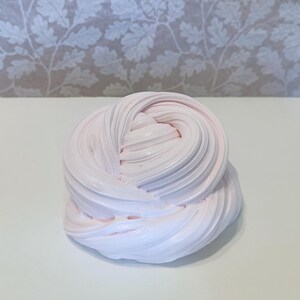 May include: A swirl of light pink slime, with a smooth, textured appearance. The slime is a soft, pastel color and appears to be freshly made. The background is a white surface with a floral pattern.