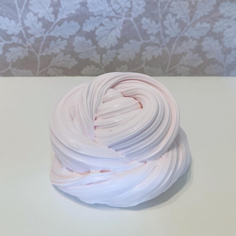 May include: A swirl of light pink slime, with a smooth, textured appearance. The slime is a soft, pastel color and appears to be freshly made. The background is a white surface with a floral pattern.