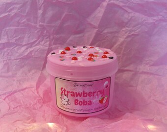 May include: Pink container of Strawberry Boba slime, decorated with red, white, and black sprinkles. The label features a cartoon cat and the text "Strawberry Boba". The background is pink tissue paper.