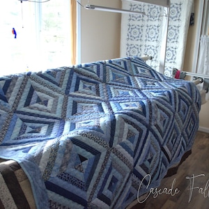 May include: A handmade quilt in shades of blue, with a geometric diamond pattern. The quilt is displayed on a frame, showcasing the intricate stitching and design. The text "Cascade Falls Quilts" is visible in the lower right corner.