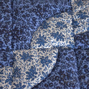 May include: Close-up of a quilted fabric with a floral pattern in shades of blue and white. The design features a combination of solid blue and patterned sections, creating a textured appearance. The quilting stitches are visible, adding to the handmade aesthetic.