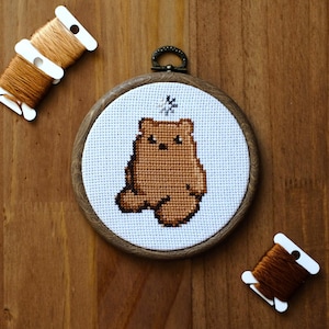 May include: A cross-stitch embroidery of a brown bear in a wooden hoop. The bear is stitched on white fabric. Three spools of brown thread are arranged on a wooden surface. A small metal ring is attached to the top of the hoop.