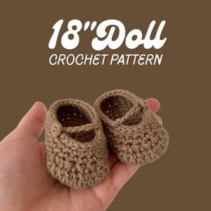 Può includere: Un paio di scarpe per bambole lavorate all'uncinetto in marrone con un cinturino e un piccolo bottone su ogni lato. Il testo 18" Doll CROCHET PATTERN è visualizzato sopra le scarpe. Presentano una trama a maglia visibile e punta arrotondata.