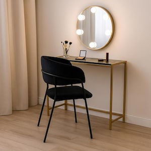 May include: A black vanity chair and a gold-framed vanity table with a marble top. A round mirror with lights is mounted above the table. Makeup brushes and products are on the table.